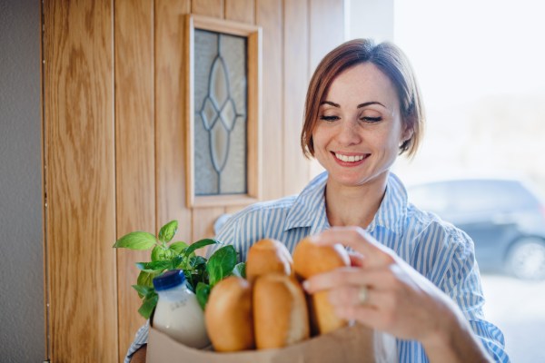 A young woman with groceries in paper shopping bag walking in through front door.