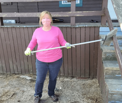 A middle aged woman wearing jeans and a hot pink shirt stands in front of a YMCA camp cabin and sweeps cobwebs from a stair rail.