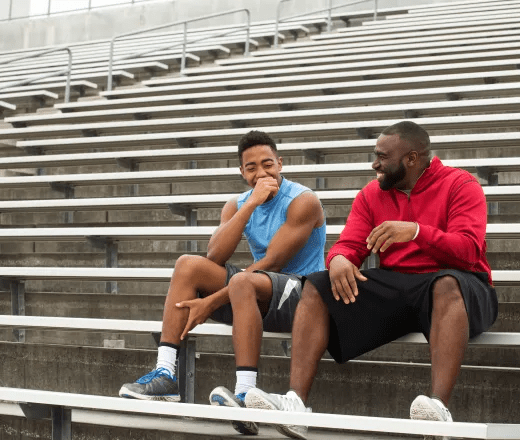 Two men, one younger and one older wearing athletic wear sit on bleachers laughing and talking.