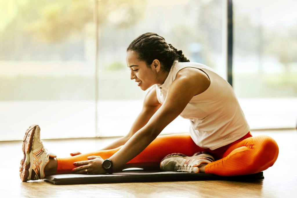 woman wearing running shoes, orange running pants, and a white sleeveless top seated on a mat on the floor stretching her hamstrings.
