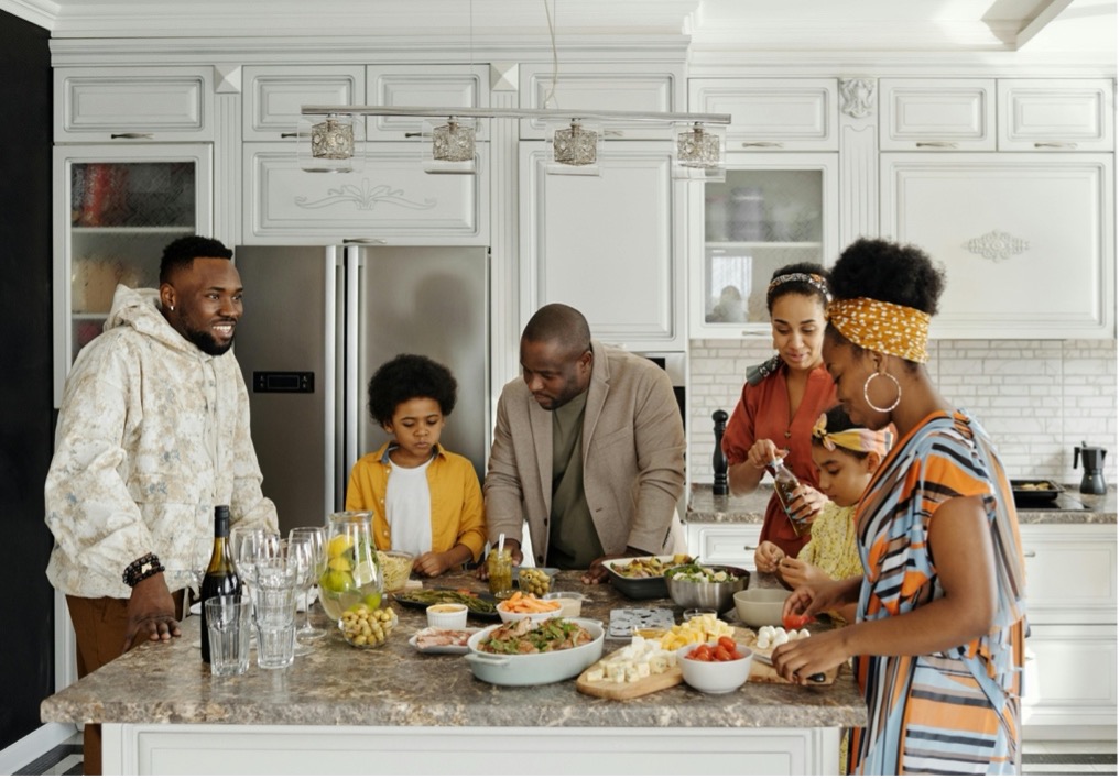 An African-American family dressed up for the holidays gathers around a kitchen island 
 that has appetizers and drinks set out on it.