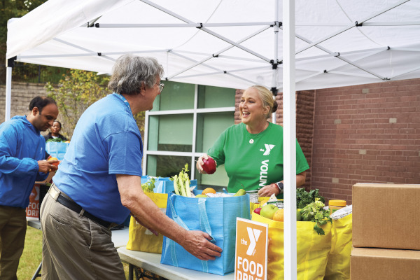 a YMCA volunteer wearing a green t-shirt places an apple into a reusable grocery bag containing  fresh produce at a YMCA food drive held outside underneath a popup canopy. The bag is being picked up by an older man wearing khakis an a bright blue t-shirt.