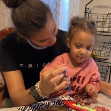 A young woman holding a girl toddler on her lap sits at a table playing together with color markers.