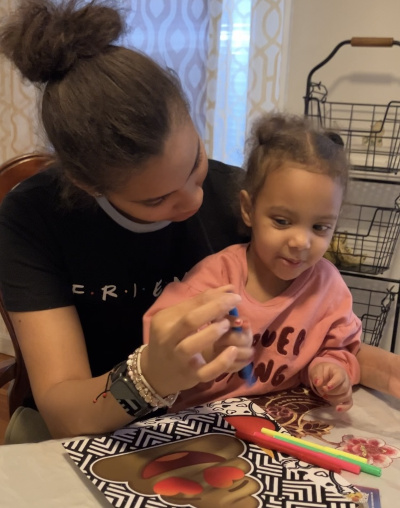 A young woman holding a girl toddler on her lap sits at a table playing together with color markers.