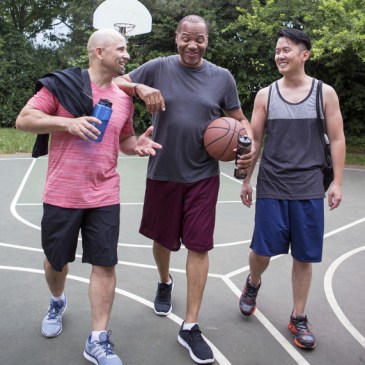 a mixed-race group of three middle-aged men, one holding a basketball, laugh with each other while walking across an outdoor basketball court looking as if they are about to play a game of basketball.