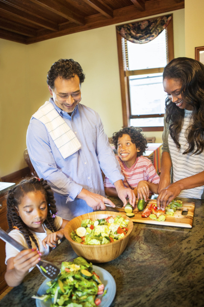 A mixed-race family (man, woman, and two children) work together at a granite kitchen counter, slicing vegetables to make a healthy garden salad.