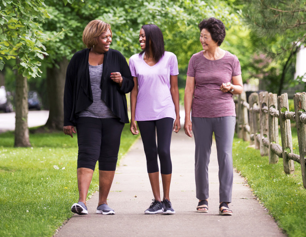 Three middle-aged women wearing exercise clothes talk with each others as they walk on paved walking path with green trees overhead and a wooden rail fence on their left.